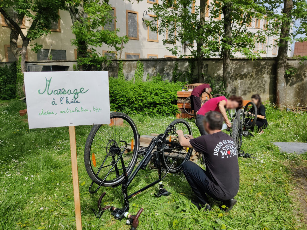 Un groupe de personnes nettoient leur chaîne de vélo dans un jardin. Au premier plan un pancarte faite à la main indique "Massage à l'huile"